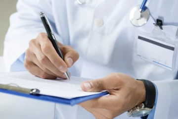 A person writing on paper with pen and stethoscope around neck.