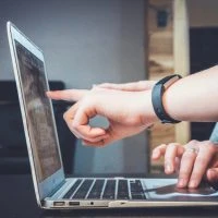 A person using their laptop with one hand and the other finger pointing at it.